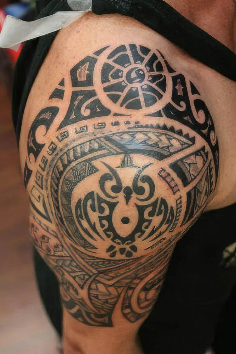 Close-up of a person's shoulder featuring a detailed Polynesian tribal tattoo with intricate geometric patterns and symbols.