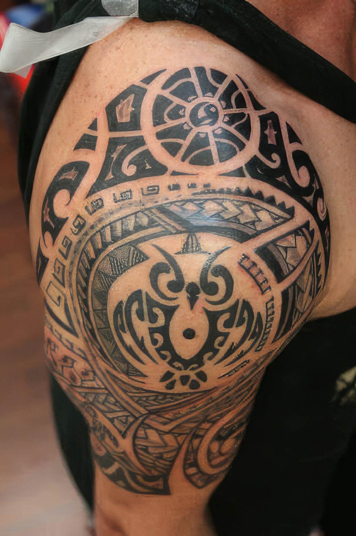 Close-up of a person's shoulder featuring a detailed Polynesian tribal tattoo with intricate geometric patterns and symbols.