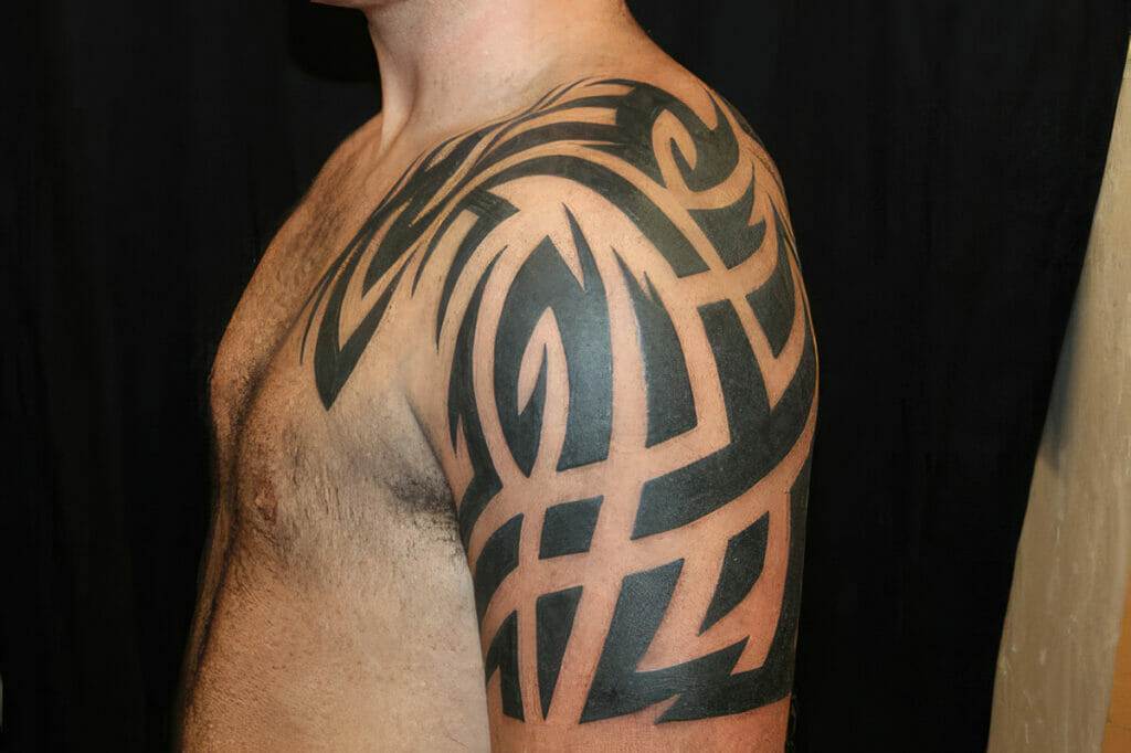 A person with a large black tribal tattoo covering their shoulder and upper arm, standing against a black background.