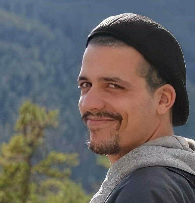 Smiling man with a beard and mustache, wearing a black cap and gray hoodie, outdoors.