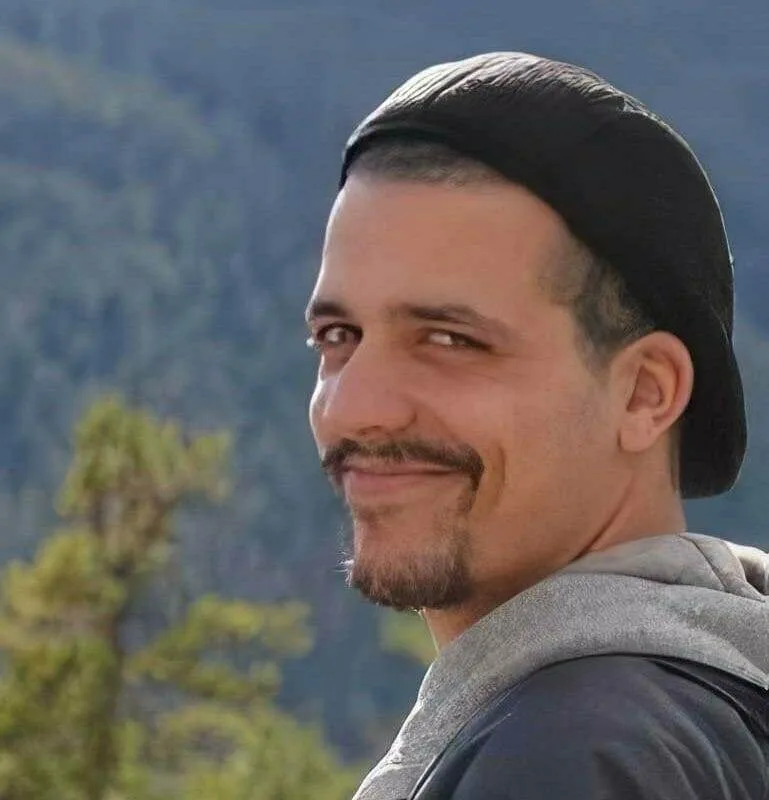 Smiling man with a beard and mustache, wearing a black cap and gray hoodie, outdoors.