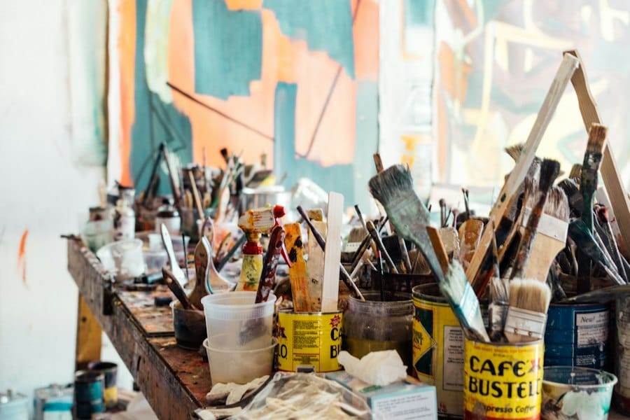 A cluttered artist's workspace with various paintbrushes, containers, and paint tubes on a table. Large canvases with abstract artwork are visible in the background.
