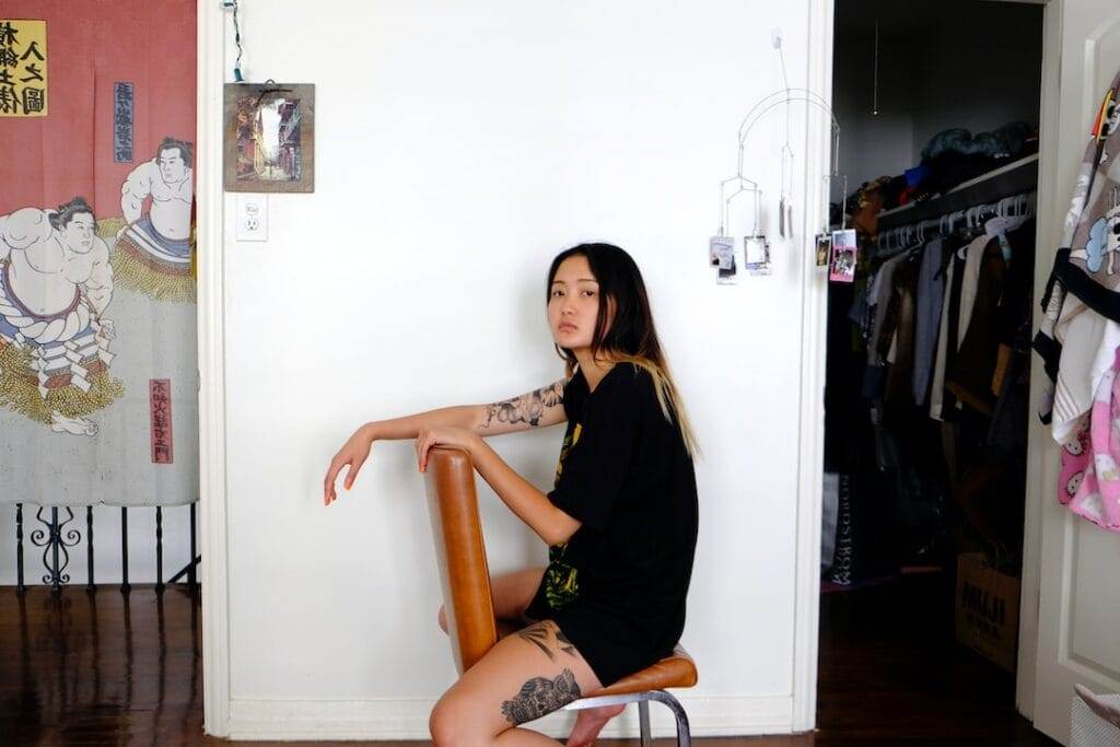 A person with tattoos sits on a chair in a room with a white wall, artwork, a closet, and various items.