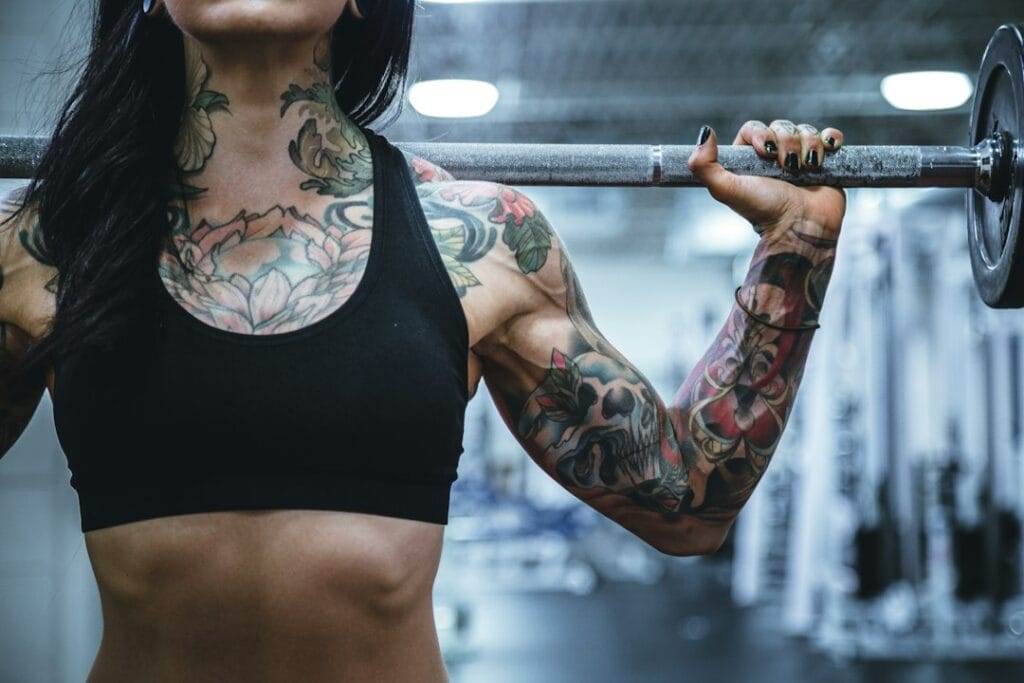 A person with tattoos on their arms and chest, wearing a black sports bra, holds a barbell across their shoulders in a gym setting.