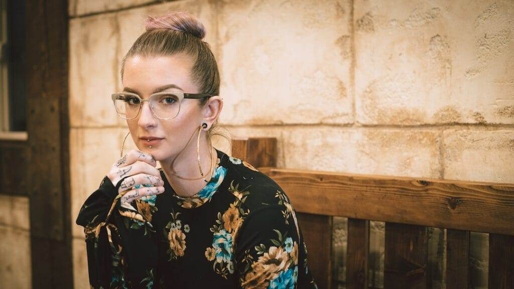 Woman with glasses and a floral top sits on a wooden bench, resting her chin on her hand. She has her hair in a bun and is looking at the camera. Background has rustic wall panels.