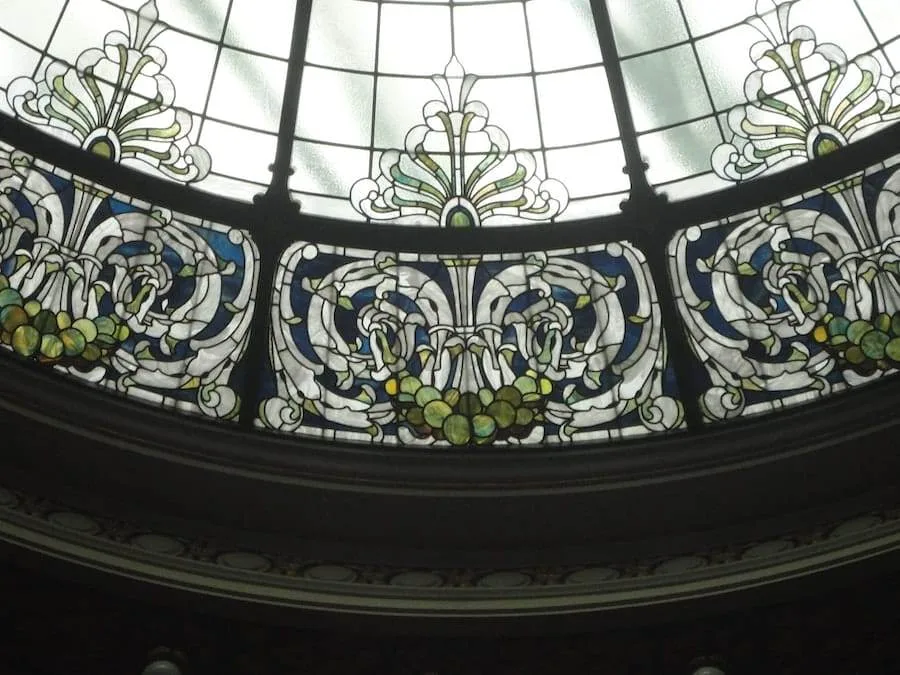 Stained glass window with intricate floral and geometric patterns, featuring green, blue, and white colors, set in a circular dome structure.