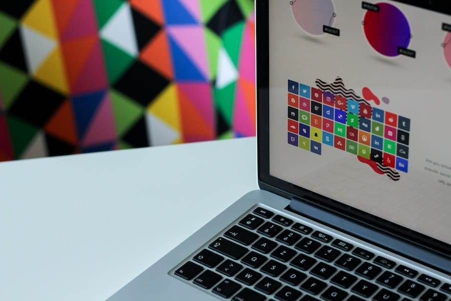 A laptop on a white desk displays a screen with colorful icons and graphics, while the background features a vibrant geometric pattern.
