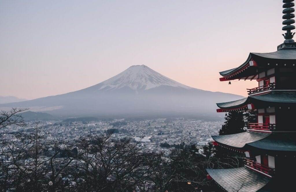 Image of Mount Fuji in the distance during twilight, with a traditional Japanese pagoda in the foreground. Mist partially covers the city at the base of the mountain.