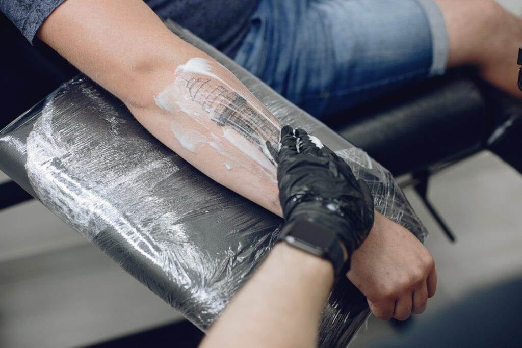 A person’s forearm is being prepped for a tattoo with cream. The person is seated, and a gloved hand is applying the cream while holding the arm steady.