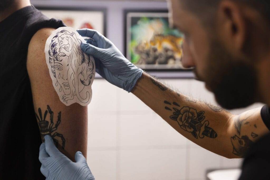 A tattoo artist applies a lion design stencil to a person's upper arm, preparing to begin a tattoo. The artist wears blue gloves, and the tattoo parlor has framed artwork on the walls.