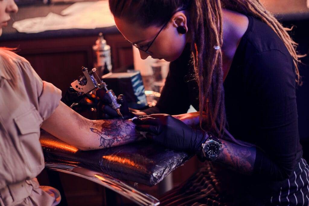 A tattoo artist with long dreadlocks is focused on creating a design on a client's forearm, using a tattoo machine. The client is seated comfortably, their arm resting on a padded surface.