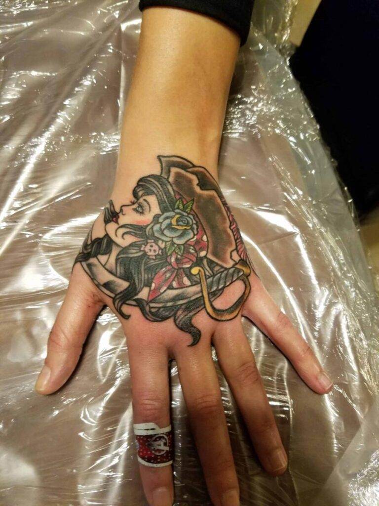 A tattoo of a woman with dark hair and red accessories is on the back of a hand that is resting on a plastic-covered surface.