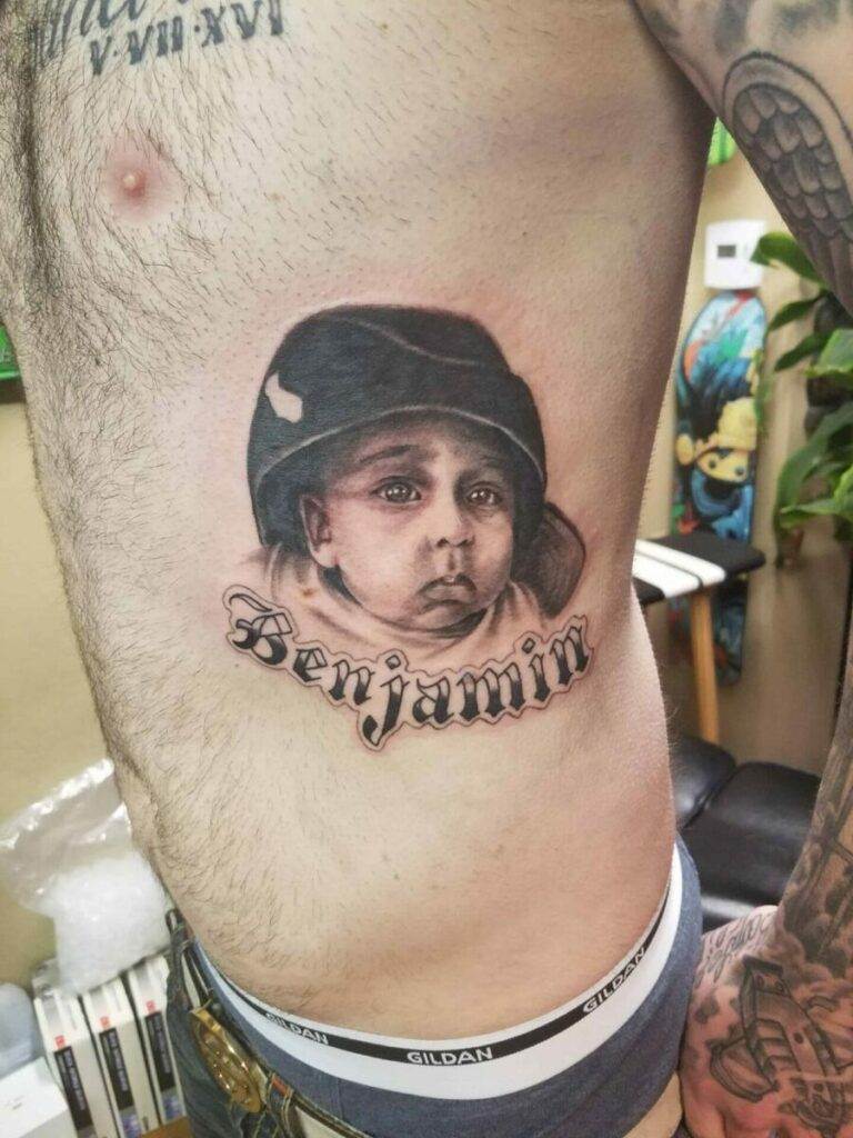 Tattoo of a child's face wearing a hat with the name "Benjamin" written underneath on a man's ribcage. There are visible chest and abdominal hair around the tattoo.