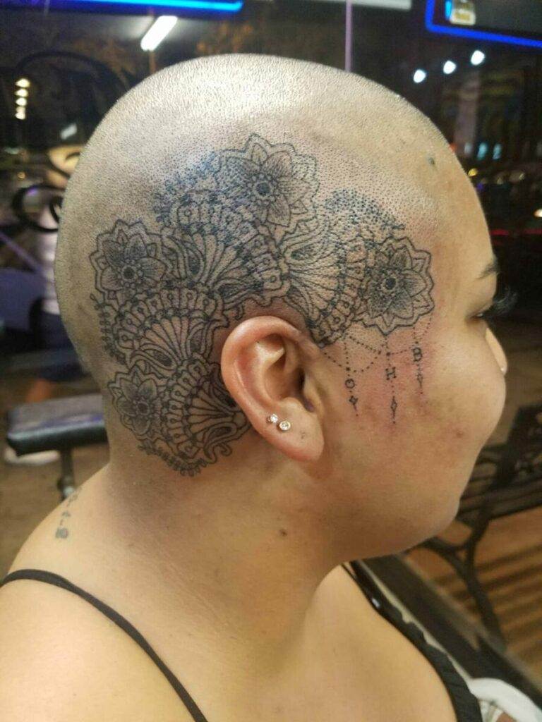 A person with a shaved head displays an intricate floral and geometric tattoo design on the side of their head.