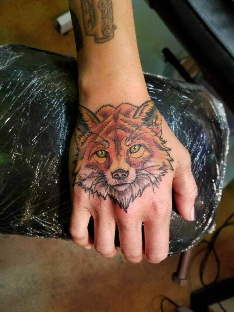 A detailed fox face tattoo on the back of a person's hand. The hand rests on a surface covered with plastic wrap.