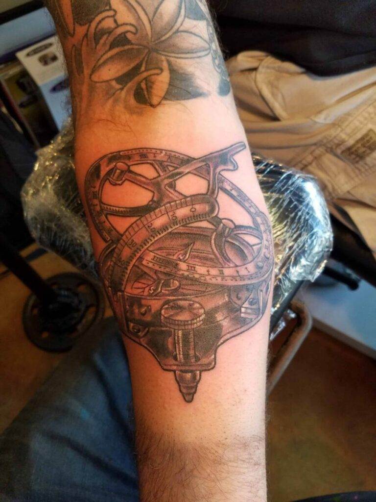 Forearm tattoo featuring a detailed, intricate design of an antique navigational instrument on a person’s arm. The tattoo is in black and gray ink.