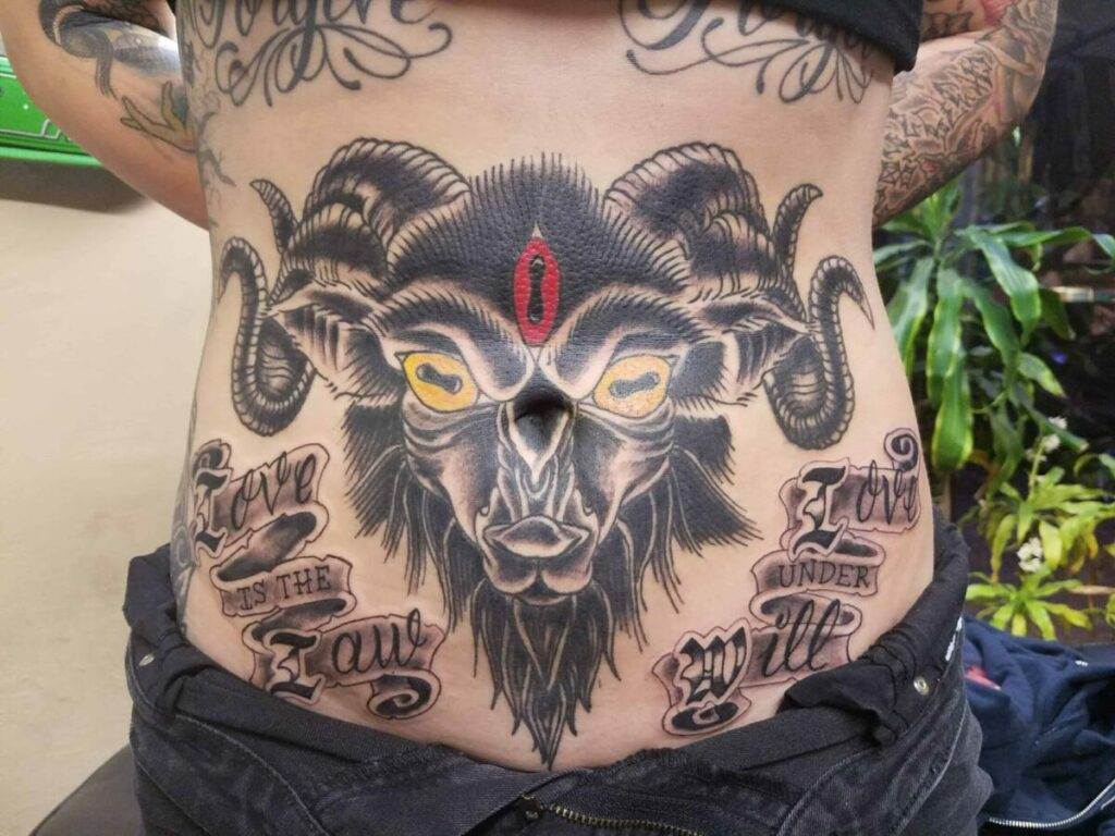 A person with a detailed tattoo of a goat with prominent horns and yellow eyes on their torso, featuring text that reads "Love is the law" and "Love under will.