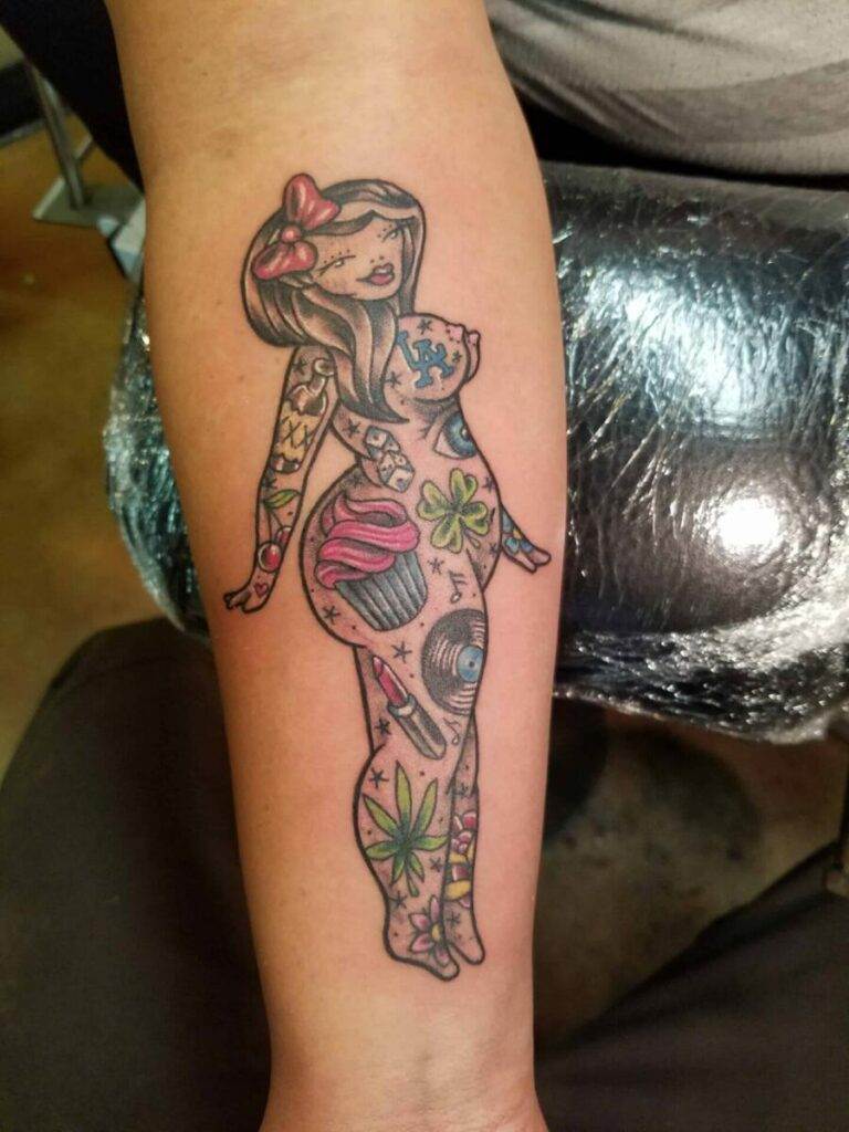 Tattoo of a cartoon-style woman covered in various colorful objects, including a cupcake, music record, and green leaves, on a person's forearm.