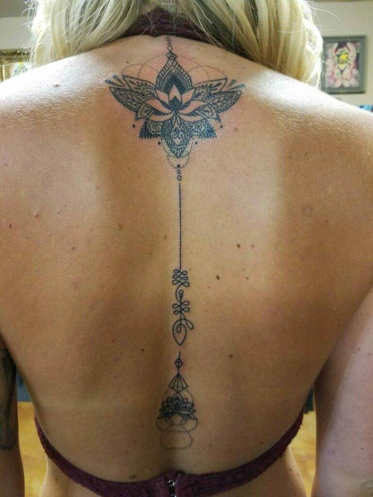 A person's back with an intricate tattoo extending from the upper back to the middle, featuring a mandala-like design at the top connected by a fine line to a smaller stylized symbol at the bottom.