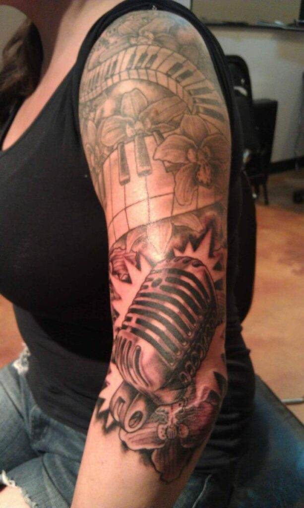 A person with a detailed black and white tattoo sleeve on their left arm, featuring a microphone, piano keys, flowers, and other intricate designs.