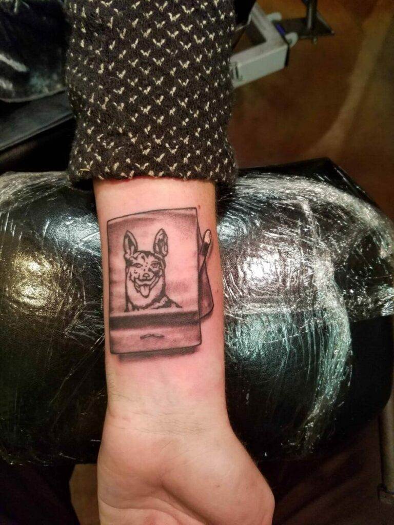 Forearm with a tattoo of a dog's head depicted in a Polaroid-style framed picture.