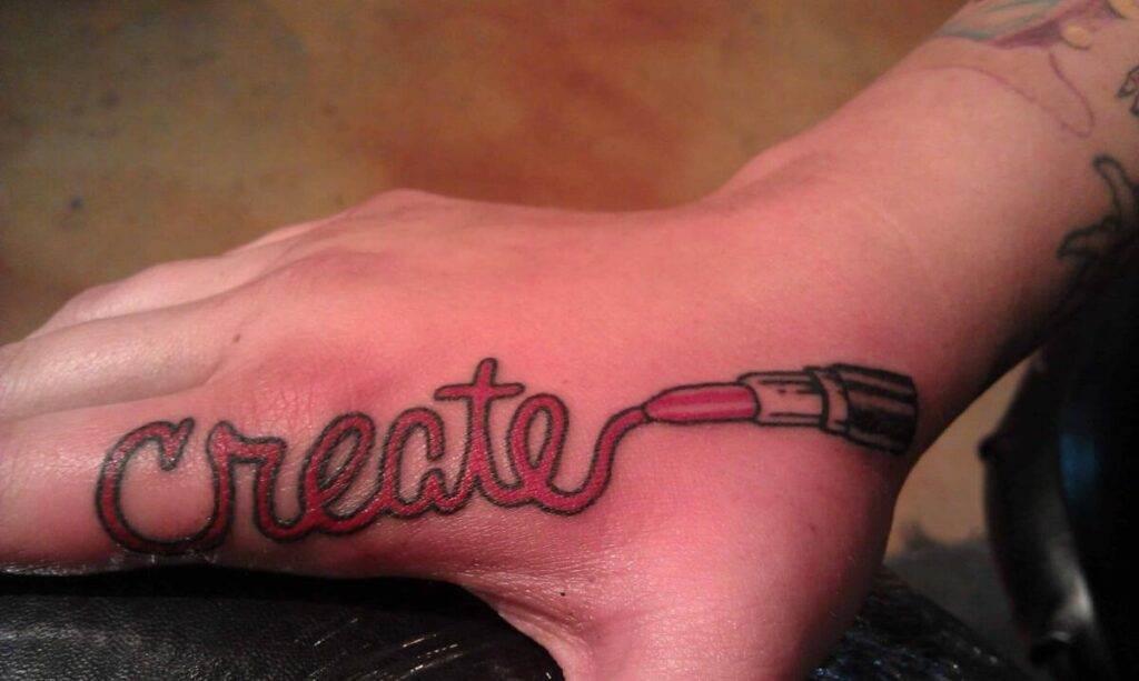 A tattoo on a hand depicting the word "create," with the tail of the last letter extending to form an image of a lipstick.