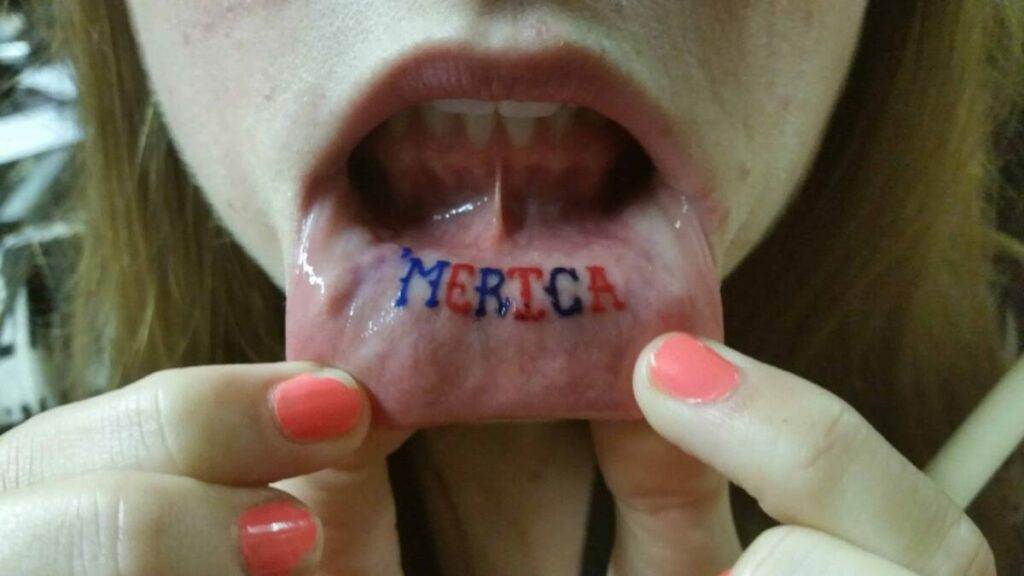 Person pulling down their lower lip to show a tattoo on the inner lip that reads "MERICA" in red and blue ink.