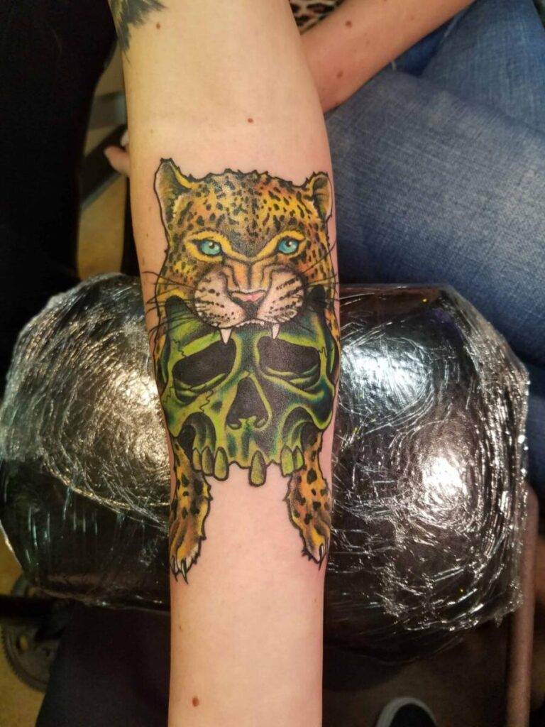 Forearm tattoo of a leopard holding a green, skull-shaped object in its mouth. Background shows a person dressed in jeans.