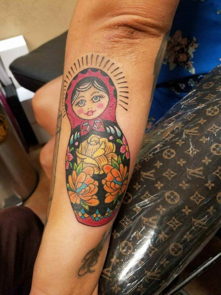 A tattoo of a brightly colored, traditional Russian matryoshka doll on a forearm, featuring floral decorations.