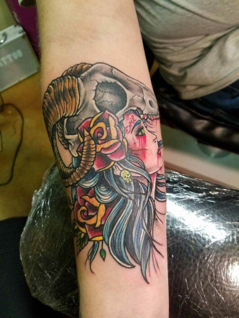 A forearm tattoo featuring a woman with blue hair, surrounded by yellow and orange roses, beneath a ram skull with horns.