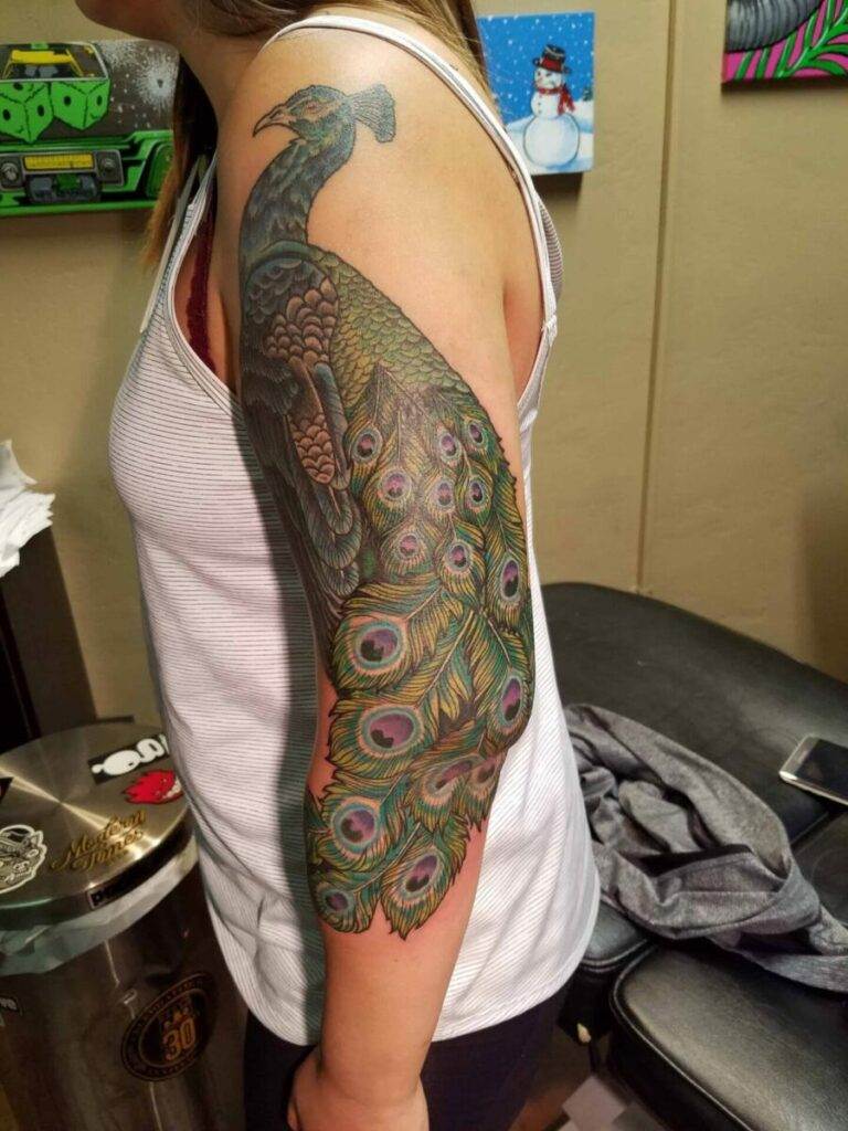 A detailed peacock tattoo covers the upper arm of a person wearing a white tank top. The vibrant design features intricate feathers and extends from the shoulder to the mid-forearm.