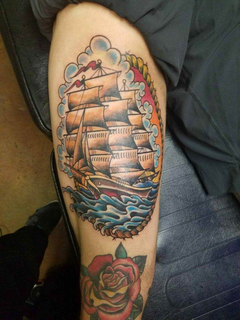 Colorful tattoo on a person's calf depicting a sailing ship surrounded by a swirling sea and clouds; a rose tattoo is visible on the ankle.