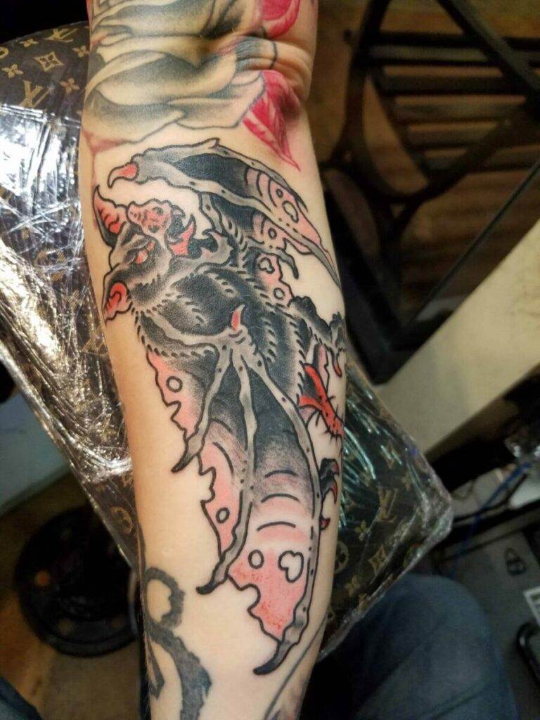 An arm features a detailed tattoo of a bat with wings spread, accented with red and pink hues.