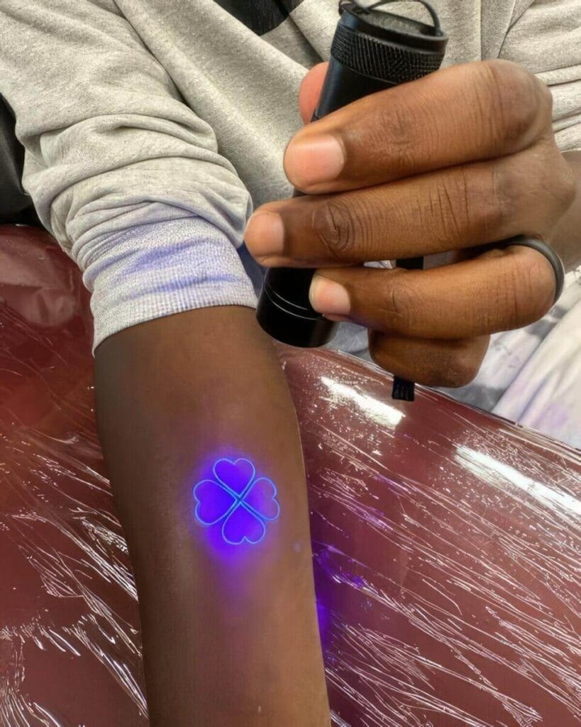 A person holds a UV flashlight over an arm, revealing a glowing design of two clover leaves, forming a heart shape.