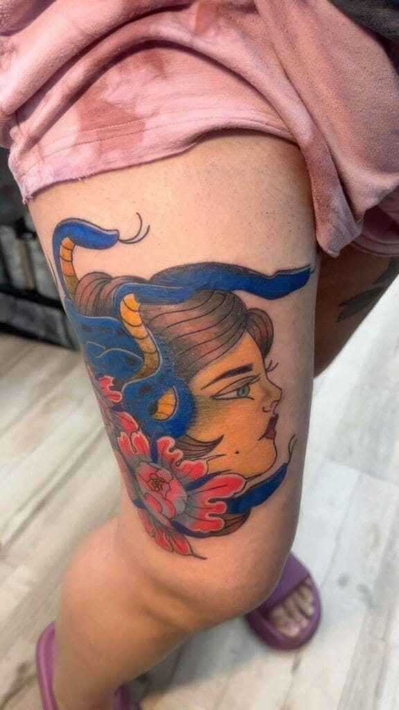 A colorful tattoo of a woman's face with blue hair, horns, and a red and blue flower is depicted on a person's upper thigh. The person is wearing pink shorts and purple footwear.
