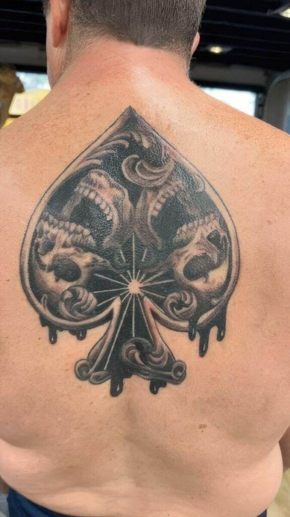 A man displays a large spade-shaped tattoo on his back, featuring intricate designs of skulls and dripping effects.