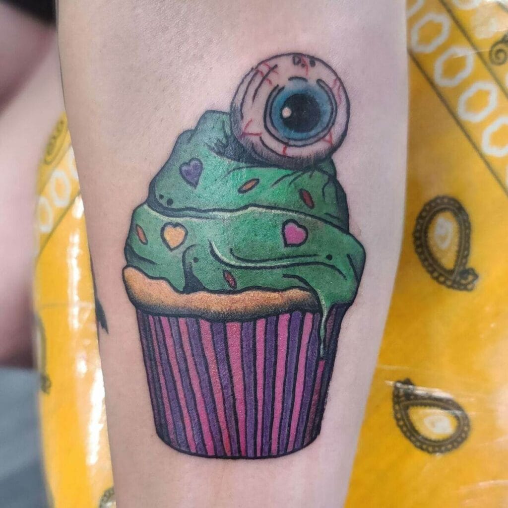 A tattoo of a cupcake with green frosting, small heart decorations, and a large realistic eyeball on top.