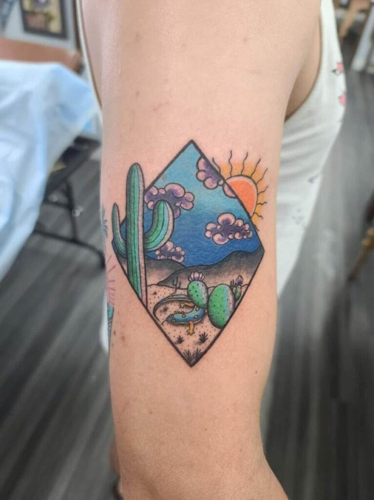 A colorful tattoo on an upper arm depicting a desert scene with cacti, pink clouds, and a setting sun.
