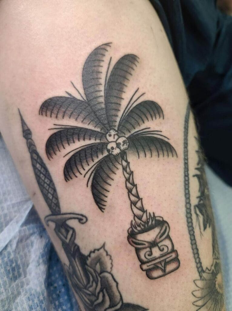 A black and grey tattoo of a palm tree with coconuts and a tiki mask is inked on a person’s skin.