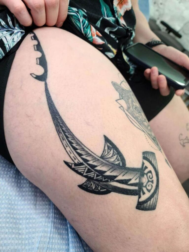 Person's thigh with a large black tribal tattoo of a stylized hook, featuring intricate patterns. The person holds a dark object in their hand and wears patterned clothing.