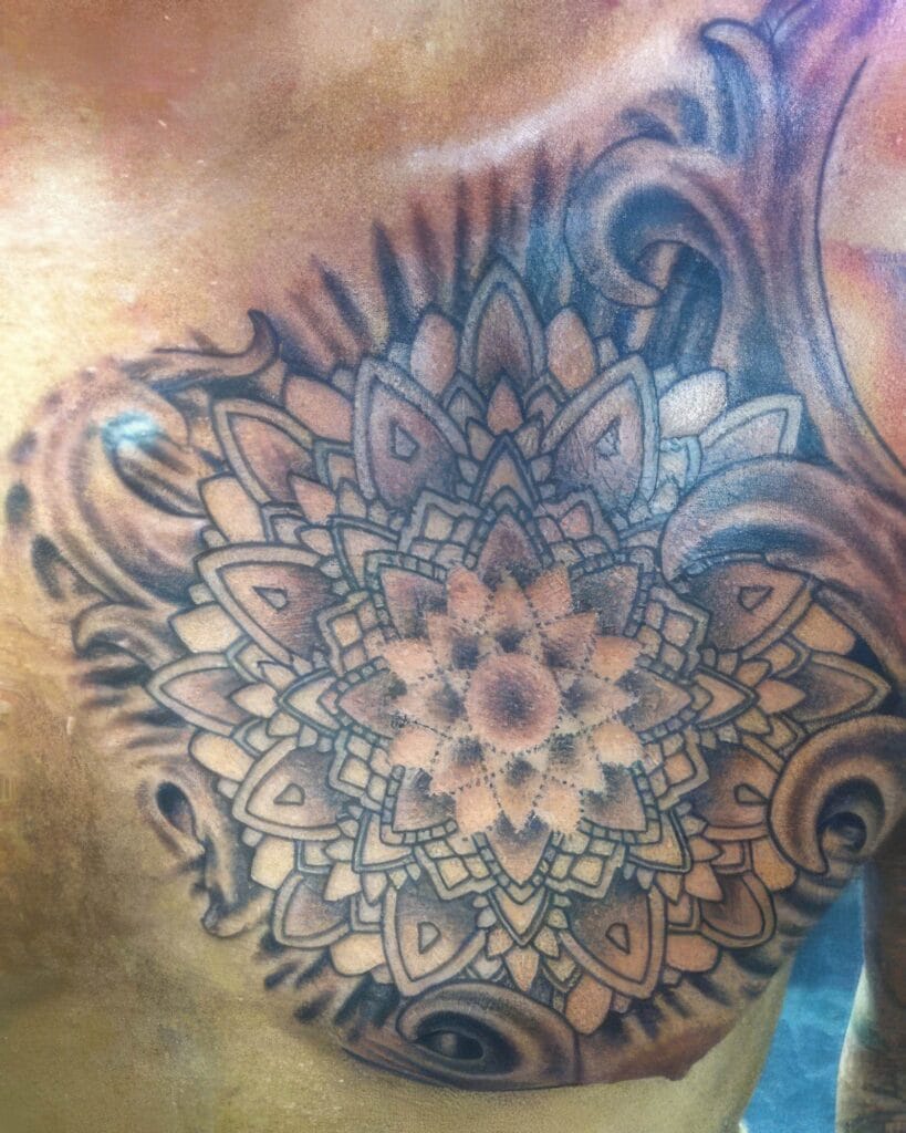 A detailed, symmetrical mandala tattoo in black and gray ink covers a person's chest, featuring intricate patterns and radiating designs.