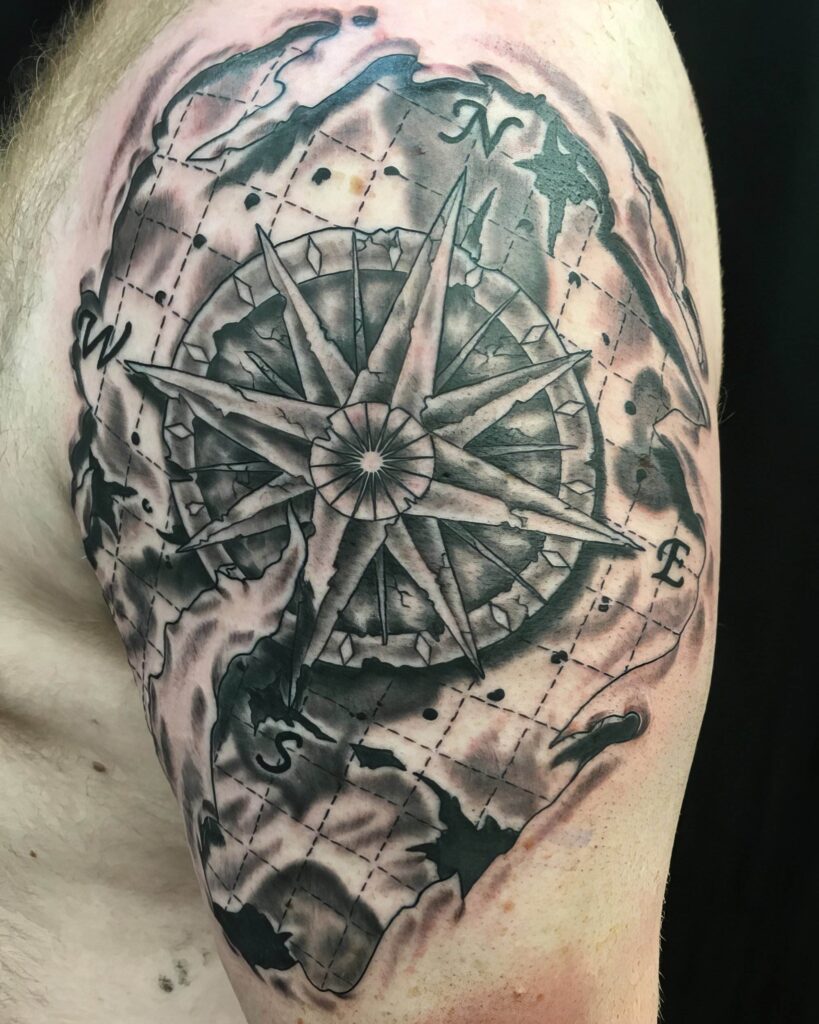 A tattoo featuring a detailed compass rose surrounded by a shaded, abstract background, located on an individual's upper arm.