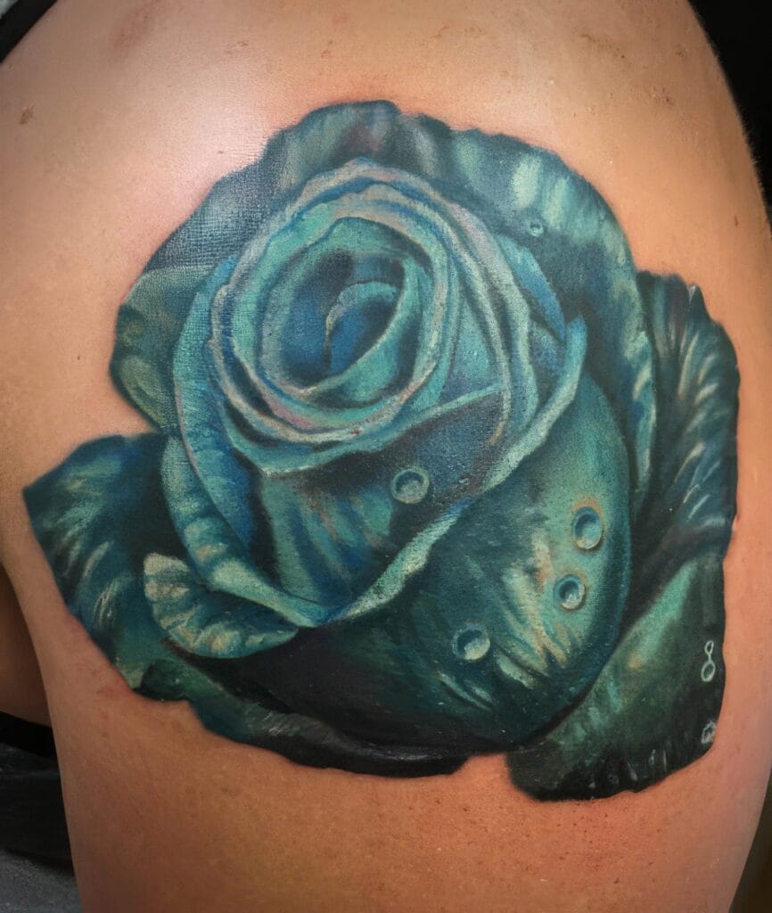 A realistic, colorful tattoo of a blue rose with water droplets on its petals, located on a person's upper arm.