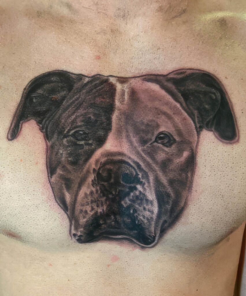 A realistic tattoo of a dog's face, predominantly shaded in black and gray, is seen on a person's chest.