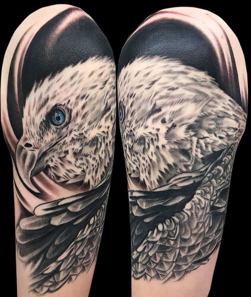 A detailed black and grey tattoo of an eagle's head and wing, positioned on a person's upper arm. The eagle's eye is blue, and its feathers are intricately shaded.