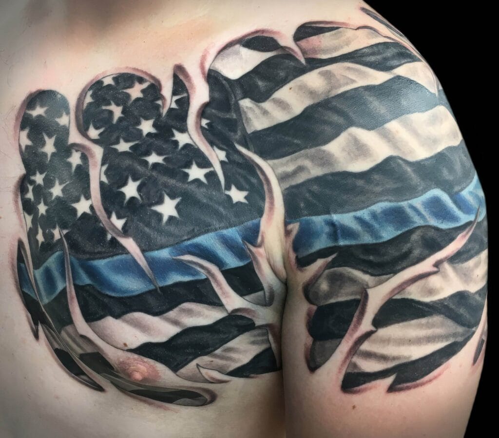 A shoulder tattoo depicts a stylized American flag with a thin blue line, symbolizing support for law enforcement. The design includes rippling effects and abstract shapes.