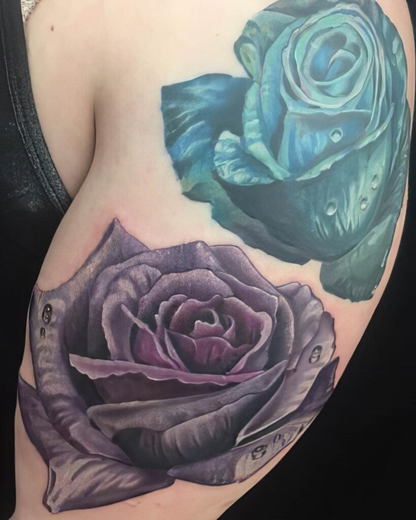 A photo showing a person's upper arm with two realistic flower tattoos, one purple and one blue. The tattoos are detailed with shading and highlights, creating a three-dimensional effect.