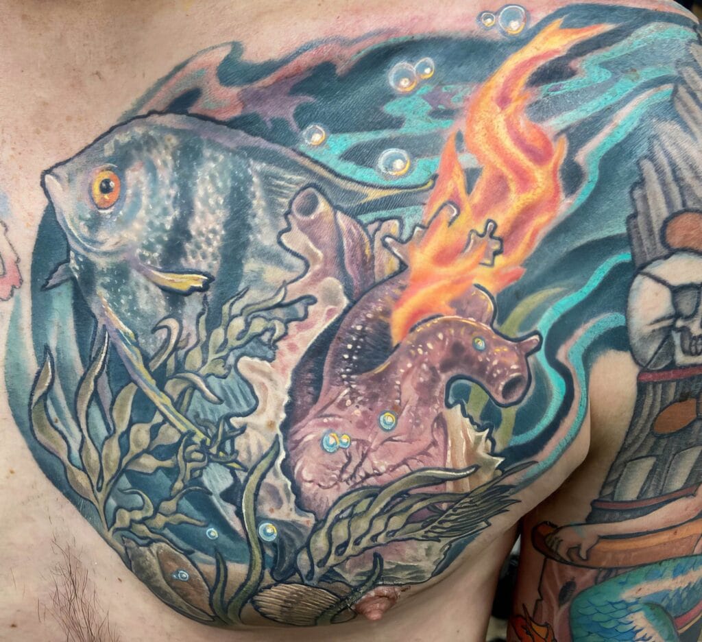 Close-up of a colorful chest tattoo featuring an underwater scene with fish, seaweed, a seashell, and a vibrant corroded object blending into waves with a fiery detail.