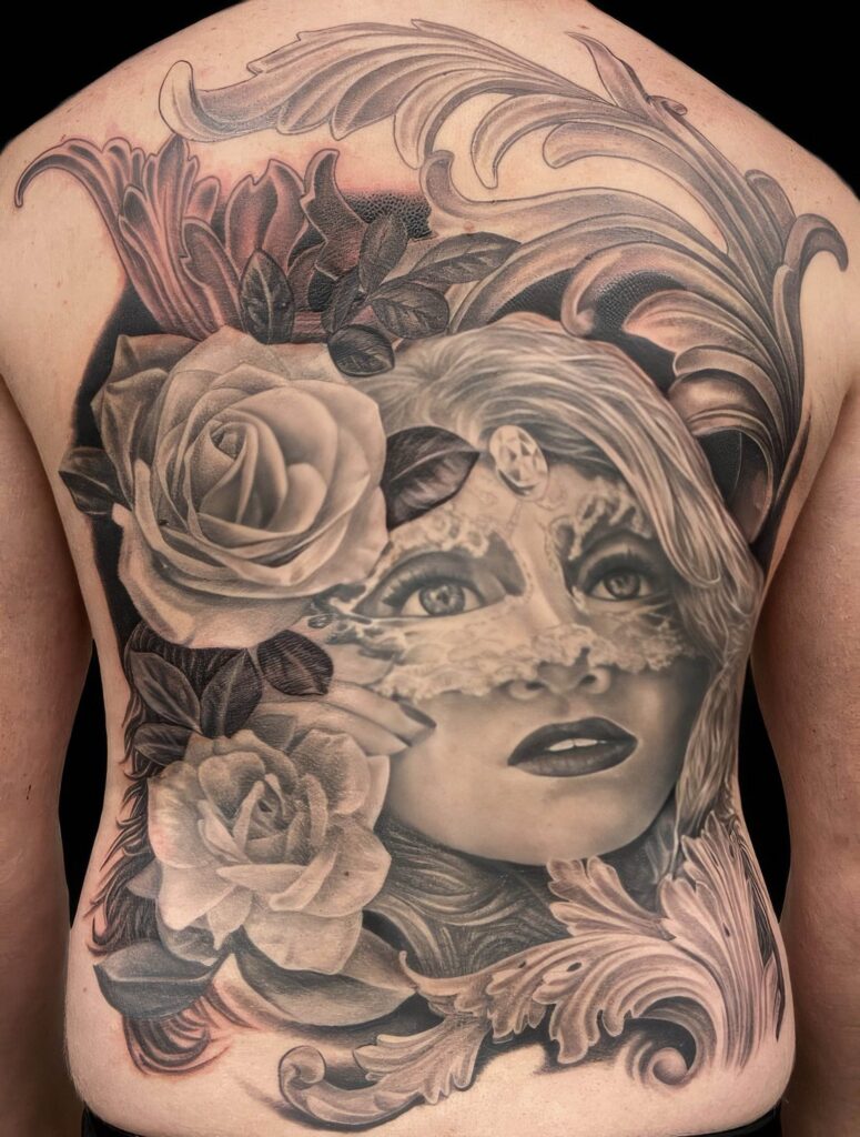 A black and gray tattoo covering the upper and middle back features an intricate design of a woman's face partially veiled with lace, surrounded by large roses and ornate patterns.