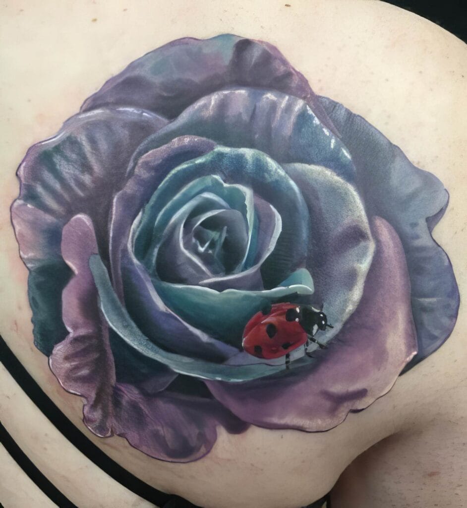 Realistic tattoo of a purple rose with a small red ladybug on its petals, located on a person's back shoulder.